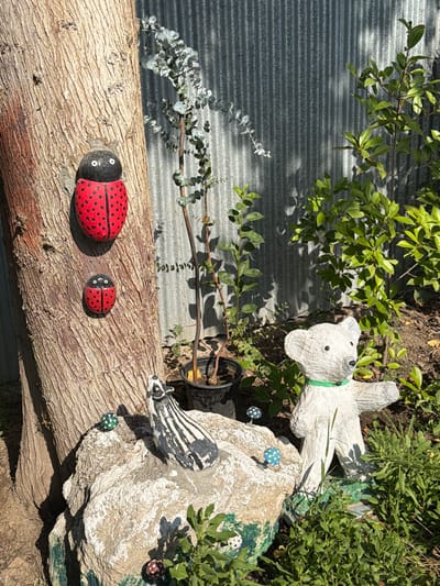 This is a close-up, eye-level outdoor shot of a garden scene. In the foreground, a large, textured tree trunk dominates the left side of the frame. Two ladybug decorations, painted red with black spots and black heads with white eyes, are attached to the trunk. The upper ladybug is larger than the lower one. To the right of the tree, a large grey corrugated metal fence forms the background. In front of the fence, a potted eucalyptus plant with sparse, grey-green leaves is visible. To the right of the eucalyptus, a white bear statue with a green collar stands among lush green foliage. In the mid-ground, a rough, light-colored rock is situated in front of the tree trunk. On the rock, a black and white striped animal figurine, possibly a skunk or badger, is placed. Several small, decorative mushrooms with white caps and blue or red spots are scattered around the rock. The overall lighting suggests a sunny day, casting shadows from the plants onto the fence. The location is identified as Balakənd, Azerbaijan.