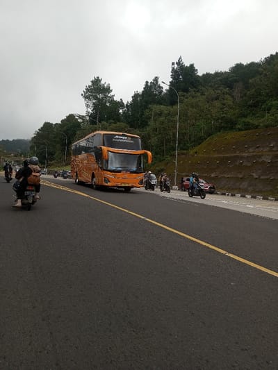 The image displays an overcast daytime scene on an asphalt road in Saing Ta Mawt Lay, Myanmar. A prominent orange high-decker passenger bus, branded "JETBUS 3" and "JAKARTA RAYA," is centrally positioned, traveling towards the viewer. A dark-colored motorcycle, occupied by two individuals, one wearing a backpack, is visible in the foreground on the left, moving away from the camera; its license plate reads B 4583 PZT. Several other motorcycles, each carrying one or two riders, are distributed around the bus, some appearing on the right lane or paved shoulder. A red passenger car is partially visible on the far right shoulder, obscured by a motorcycle. The road features a solid yellow center line and white edge lines on the right. To the immediate right of the road, a grey utility pole with a street light is present. The landscape on both sides of the road consists of ascending terrain; the left is densely forested with green trees, while the right exhibits a terraced hillside with sparse green vegetation and exposed soil.