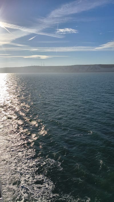 The image captures a sunlit view of the sea off the coast of Dover, United Kingdom. The sea is a deep blue-green, with sunlight reflecting off the choppy surface, creating a shimmering effect. In the distance, a white cliff face, characteristic of Dover's coastline, is visible, topped with a faint haze. On the cliff, several structures, including what appear to be radio towers or pylons, are discernible. The sky above is a bright blue, with scattered white clouds and contrails from aircraft. The foreground shows a white trail of sea foam, suggesting movement, likely from a vessel. The overall scene is natural and coastal, dominated by the sea, sky, and cliffs.
