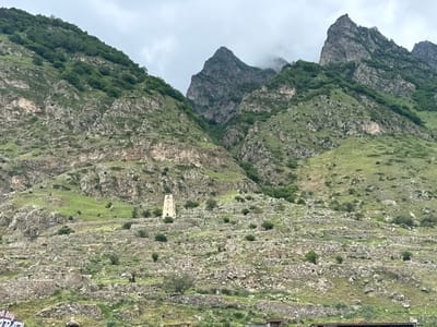 The image depicts a rugged mountain landscape in Verkhnyaya Balkariya, Russia. The scene is dominated by steep, rocky slopes covered in patches of green vegetation and trees, indicative of a summer or late spring day. The sky is overcast with clouds, suggesting a cool or potentially rainy atmosphere.

In the middle ground, a prominent, tall, stone watchtower stands on a terraced hillside. The terraces themselves are constructed from numerous stones, suggesting ancient human settlement or agricultural activity. Scattered across the slopes are the ruins of stone structures, further emphasizing the historical nature of the site. The overall impression is one of a remote, mountainous region with a rich, ancient past.

No people are clearly visible in the image, though the presence of paths and the ruined structures hint at past human habitation. The focus is on the natural grandeur of the mountains and the remnants of human presence within this dramatic setting. The lighting is diffused due to the cloud cover, casting a somewhat subdued and dramatic mood over the landscape. There is no visible text in the image.