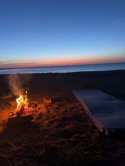 A nighttime scene of a beach campsite on the coast of Thanet, United Kingdom. The sky transitions from a deep blue overhead to hues of orange and pink near the horizon, indicating twilight or dawn. A small bonfire burns brightly in the foreground, with logs smoldering and flames reaching upwards, emitting a visible trail of smoke. To the right of the fire, a deflated sleeping mat lies on the sandy ground, partially covered with dried grass and twigs. The sea is visible in the background, appearing calm and reflecting the faint light of the sky. The overall atmosphere suggests a solitary and peaceful camping experience.