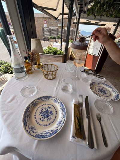 This image captures a table setting on an outdoor patio, likely at a restaurant in Saint-Tropez, France. The sun is shining, suggesting it's daytime, and the warm weather is ideal for al fresco dining. The table is covered with a crisp white tablecloth, adorned with blue and white patterned plates, silverware, and glassware. A person's hand is visible in the upper left, holding a spoon over a wooden menu holder with the word "Bou" printed on it.

Among the objects on the table are several glasses, one of which appears to be a wine glass filled with a light-colored beverage. There's also a glass bottle filled with a clear liquid, possibly water, and a small amber-colored bottle that might contain a condiment or drink. A decorative gold wire basket stands near a lamp with a pleated shade and a dried floral arrangement.

A distinctive detail is a small, individually wrapped refreshment towel placed on top of a napkin next to the silverware. The text on the towel is clearly visible: "POU - RINCE-DOIGTS RAFRAICHISSANTS - REFRESHING TOWEL" and "ALOE VERA GREEN". Another napkin also has "Bou" printed on it.

In the background, through an open frame, glimpses of a picturesque street with stone buildings and a bicycle suggest the restaurant's location. The overall atmosphere is one of relaxed dining and enjoyment of good weather.