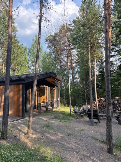 The image depicts a modern wooden cabin nestled within a pine forest. The cabin features dark wooden siding and large windows that reflect the surrounding trees and sky. In front of the cabin, there is a gravel path leading to wooden lounge chairs stacked against a pile of chopped firewood. The scene is illuminated by natural sunlight, suggesting it is daytime. The sky is partly cloudy. The surrounding trees are tall and slender with green needles, characteristic of a northern forest. The overall impression is one of a tranquil, rustic retreat in a natural setting. There is no visible text in the image.