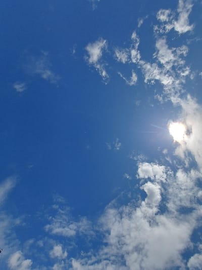The image is a daytime shot of a blue sky with white clouds, taken at the coordinates 49.211, -122.682, which corresponds to a location in Pitt Meadows, Canada. The sun is shining brightly, with its glare visible in the top right corner of the frame. The clouds are scattered throughout the sky, with some appearing more dense than others. There are no people or objects visible in the image.