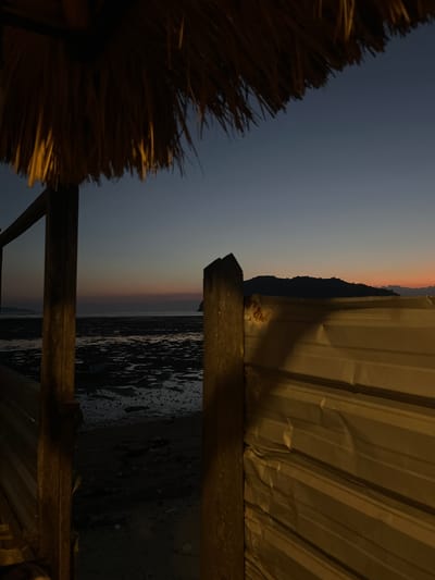 The image depicts a twilight scene on the beach in Kertasari, Indonesia, viewed from under a thatched roof structure. In the foreground, a wooden beam and corrugated metal wall partially obscure the view of the beach. The beach itself is partially submerged, with shallow water reflecting the dusky sky. A small boat is visible in the water. In the distance, a silhouette of a hilly landscape is outlined against the horizon, where the sky transitions from a deep blue to hues of orange and pink. The overall atmosphere is serene, characteristic of a coastal sunset.