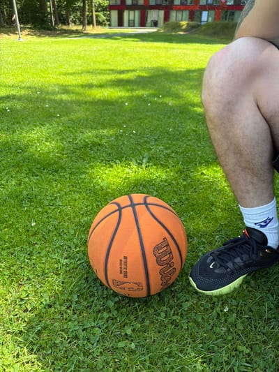 An outdoor medium shot captures a close-up view of an orange basketball positioned on a vibrant green lawn. To the right of the basketball, a person's right leg and foot are visible, clad in a black athletic shoe with a neon green sole and white socks. The background features a modern building with red and dark brown sections, partially obscured by trees. The scene appears to be a park or recreational area, possibly within the city of Enschede, Netherlands, given the presence of the modern architecture and manicured green space. The lighting suggests it is daytime, with shadows cast by unseen objects indicating a sunny or partly cloudy sky.