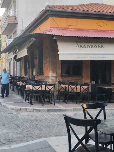 This image depicts the exterior of a restaurant in Alexandroupolis, Greece, specifically at "Η ΑΓΟΡΑ ΛΑΔΟΚΟΛΑ" located at THΛ: 25510-80701. The establishment features an orange facade with a red corrugated metal roof, and an awning extending over the outdoor seating area. Black chairs and tables are arranged on the cobblestone sidewalk, with some tables covered in brown paper. A lone elderly man with a blue shirt and dark pants is walking past the establishment. The scene is framed by buildings and street elements, including cobblestone pavement, a sidewalk curb, and a small drain grate on the ground. The overall impression is of a quiet street scene in a Greek city.
