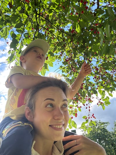 In this close-up, upside-down selfie, two individuals are positioned beneath a tree laden with bright red cherries against a backdrop of a bright blue sky with scattered white clouds.
The primary subject in the foreground is a woman looking up and smiling, her face filling a significant portion of the frame. She has light skin, a visible eyebrow, and a bright smile revealing her teeth. She is wearing a cream-colored garment over a dark blue top with embroidered detailing on the shoulder. A ring is visible on her right hand, which rests near her head.
Partially visible behind her and to the left is a young child wearing a light green cap and a yellow t-shirt with red flames. The child is also looking upwards, with their mouth slightly open. Their left arm is raised, reaching towards the cherry branches.
The scene is dominated by the lush green leaves and the red cherries of the tree, suggesting an outdoor setting during the fruit-bearing season. The upward angle of the photo, combined with the bright sky and sunshine filtering through the leaves, indicates it is daytime, likely a sunny afternoon. The overall mood appears joyful and engaged, with the subjects seemingly interacting with the cherry tree. The location context is Rezekne, Latvia. There is no visible text in the image.