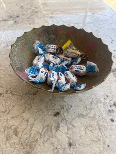 A close-up shot shows a decorative, ornate, brass-colored bowl filled with individually wrapped candies. The candies are primarily white with blue and white checkered patterns on the wrappers, and some have text such as "MILKY SPLASH TOFFEE" and "ROSHEN" visible. There are also a few candies with brown and gold wrappers. The bowl has intricate etched designs and appears to be sitting on a speckled, light-colored countertop, possibly granite, with a marble-like pattern. The lighting suggests an indoor setting, likely during the day. The image does not provide any information or context related to the city of Ghukasavan, Armenia.