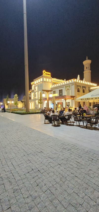 The image shows a nighttime scene outside a building labeled "HOTEL OLDHOUSE." The building is brightly lit with yellow lights, and features a prominent minaret-like structure on the left. In front of the hotel, a paved outdoor seating area is occupied by several people sitting at tables and chairs, engaged in conversation or dining. The ground is paved with stone tiles. The sky is dark, indicating it is nighttime. The overall atmosphere appears relaxed and social.