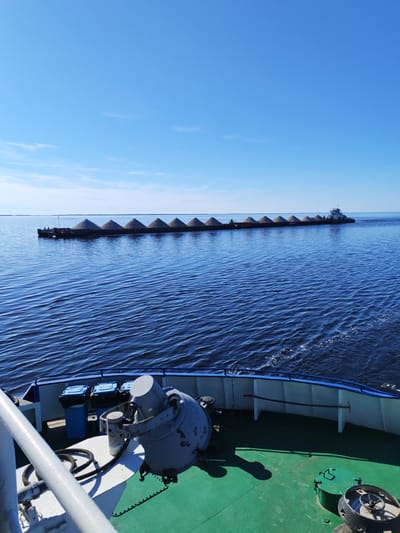 The image depicts a clear-day scene on a wide body of water, likely a river or large lake, in Vasil'evskoe, Russia, viewed from the bow of a moving vessel. In the midground, a long train of approximately 12 or more barges, each laden with a conical pile of light-colored bulk material, is being propelled from its stern by a white and blue tugboat with a multi-story superstructure. The tugboat generates a noticeable white wake in the dark blue water. The water exhibits ripples across its surface. In the foreground, the green deck of the viewer's vessel is visible, featuring various maritime equipment: a large gray winching mechanism with a coiled black rope, a white safety railing, two blue trash bins with black liners, a circular green hatch cover, and another piece of machinery with a wheel. A long pole or hook tool rests against a white bulkhead on the starboard side of the deck. Shadows cast by the equipment indicate bright sunlight. The background consists of a clear, bright blue sky with sparse, wispy clouds near the horizon, above a faint, distant landmass. No people are visible.