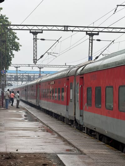 In Ballabgarh, India, a red and gray train is stopped at a platform. The train has multiple cars with visible windows. Several people are standing on the wet platform, some near the train. Overhead, electrical wires and support structures for an overhead catenary system are visible against a cloudy sky. The platform shows puddles of water and some debris. A blue sign with white text "1501 20" is attached to a pole near the train. The environment appears to be an outdoor railway station.