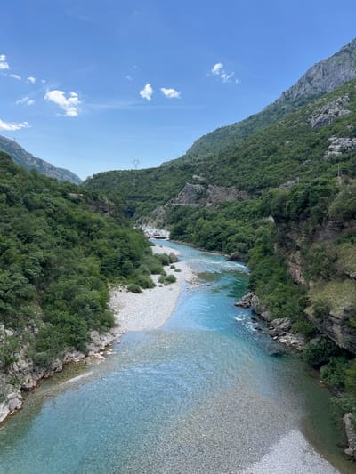 The image depicts a river flowing through a canyon in Unknown, Montenegro. The river is clear and turquoise in color, and its bed is composed of pebbles and rocks. Lush green vegetation covers the steep canyon walls. The sky is blue with scattered white clouds. Power lines are visible running along the side of the canyon.