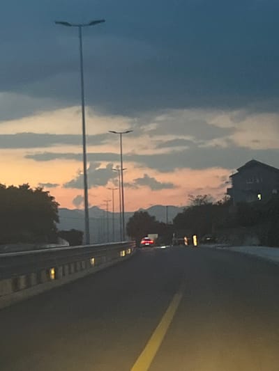 This media file depicts a view from a vehicle moving along a road at dusk. The sky is filled with clouds, displaying a gradient of blue, gray, and soft orange hues, suggesting either sunrise or sunset. In the background, a dark silhouette of a building, possibly a house, is visible against the sky. Trees line the side of the road, also appearing dark in the low light.
On the left side of the image, a street lamp with a horizontal bar is prominent, and further to the right, a series of similar street lamps are visible, casting subtle reflections. The road itself is dark asphalt with a bright yellow line marking its edge. A metal railing runs along the right side of the road, with small, illuminated lights visible along its length. There are no visible people in the frame.
The overall mood is serene and contemplative, characteristic of twilight. The scene is likely a suburban or rural area in Radanovići, Montenegro, given the context. No visible text is present in the image. The activity taking place is vehicular travel on a road during the transition from day to night.