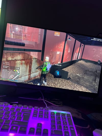 The image shows a computer monitor displaying a video game scene, with a purple-lit keyboard in the foreground. The game appears to be a first-person shooter or action-adventure game, possibly set in a gritty, industrial urban environment.
In the game scene, a player character, wearing a green jacket, blue backpack, and white cap, is visible holding a shotgun. Another player character in a blue and black outfit is seated on the ground. The environment features corrugated metal walls, wooden structures, and a large pile of what appears to be wood chips or debris. There are also in-game text overlays, indicating player names, game status, and chat messages.
The location in the city of Tampere, Finland, is not depicted in this image. The image exclusively shows a digital representation within a video game.