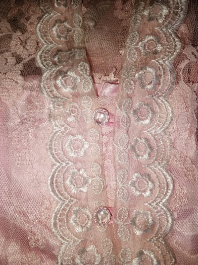 The image, taken in Na Sang, Laos, presents a detailed close-up of a pink lace garment. A central vertical placket is visible, framed by white embroidered lace trim featuring a scalloped edge and floral patterns. Two circular, clear, or pearlescent buttons, each secured with pink thread, are present along the placket. The main fabric of the garment is a delicate, light pink lace with a textured floral design. No individuals, actions, or broader environmental features are visible; the focus is solely on the textile details.