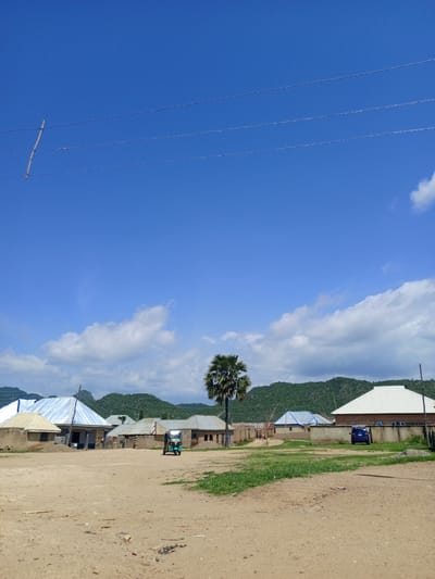 The image depicts a rural scene in Barakin Babele, Nigeria, under a clear blue sky with scattered white clouds. The time of day appears to be daytime, likely late morning or early afternoon, given the bright sunlight and the shadows cast. The weather seems pleasant and warm.
The foreground is a dirt road or open area, with scattered debris. In the mid-ground, several buildings with corrugated metal roofs of various colors are visible, indicating a small settlement or village. Some of these buildings are simple, traditional structures, while others appear more modern. A few people can be seen walking near some of the buildings. A small, colorful three-wheeled vehicle, commonly known as a "keke napep" or "Marwa," is parked in the middle of the scene. To the right, a blue SUV is parked next to a building with a white roof, separated by a concrete wall. A tall palm tree stands prominently on the left side of the image.
In the background, lush green hills rise, suggesting the village is situated in a hilly or mountainous region. Power lines are visible stretching across the upper part of the frame, supported by a wooden pole, indicating the presence of electricity in the area.
There is no visible text in the image. The overall mood is serene and peaceful, capturing a slice of everyday life in a Nigerian village.