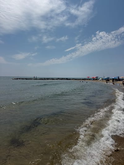 This image captures a sunny day at the beach in San Felice Circeo, Italy. The foreground shows the gentle waves of the sea lapping onto a sandy shore. In the midground, the calm water extends towards a man-made breakwater constructed of dark, irregular rocks. Two figures are visible standing on this breakwater, silhouetted against the horizon. Further down the coastline, a beach scene unfolds with several colorful umbrellas providing shade for beachgoers. People are scattered across the sand and in the shallow water. The sky above is a clear blue, punctuated by wispy white clouds. The overall scene depicts a relaxed atmosphere typical of a summer day at the seaside.