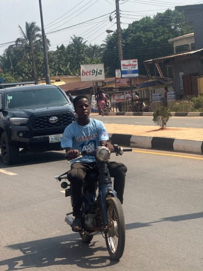 A young male operates a blue-colored, older model motorbike on an asphalt road in Aguata, Nigeria. He wears a light blue t-shirt with "HEROES 10 ATM" print, black cargo pants, and dark sandals, looking slightly to his right. His hands grip the handlebars, and his feet rest on the motorbike's frame. The motorbike features a circular headlight, spoked wheels, and exposed engine components. Behind him and to his left, a dark-colored Toyota Tacoma pickup truck with license plate "LND-863JV" is partially visible. The road has a solid white lane line on the left. To the right of the motorbike is a raised curb, painted with black and white stripes, separating the road from a dirt shoulder. A small green shrub is planted on this median. In the background, across the road, various structures are present, including a white billboard displaying "bet9ja" and "BEN GIG STREET," and a blue sign for "WoodEdge HOTEL & SUITES." Overhead utility poles and multiple power lines crisscross the scene. Several individuals are visible in the distance near the structures, including one person in a pink garment. Dense tropical vegetation, including palm trees, rises behind the buildings. Long shadows are cast on the road, indicating strong sunlight.