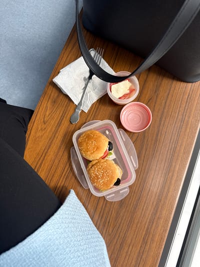 The image depicts a wooden surface, likely a table, with a lunch arrangement consisting of a black handbag, a small open container with white cheese and what appears to be strawberries in liquid, a small pink container, a small plastic container holding two sesame seed buns filled with ingredients like tomatoes and olives, a fork on a white paper napkin, and a light blue knitted fabric next to black trousers. The location is indicated as Hanau, Germany, though no specific landmarks or identifiable elements of the city are visible in the frame. There are no people explicitly depicted, but the presence of clothing and personal items suggests the photo was taken in someone's personal space or during a meal break. The environment appears indoor, evidenced by the artificial lighting and a window frame with a window pane visible at the top right of the image.