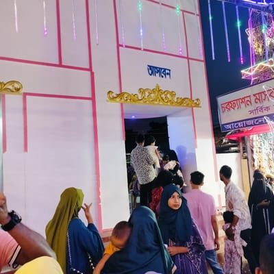 This nighttime image captures a lively scene in Charfasson, Bangladesh, with numerous people gathered around the entrance of a temporary structure. The building is white with prominent pink trim and ornate golden decorative elements above the entryway. Colorful LED string lights hang vertically, illuminating the area with a festive glow against the dark sky.

Many individuals of various ages are present, including adults and children. Several women are wearing hijabs, and one man is seen carrying a child while navigating the crowd. People appear to be moving in and out of the entrance, suggesting a public event, market, or gathering.

Visible text on the white structure above the entrance reads "আসবেন" (Asben), which translates to "Come" or "Welcome" in Bengali. To the right, a sign also in Bengali displays "চরফ্যাশন মাস্ট" (Charfasson Mast) followed by "সাবির্ক আয়োজনে" (Sabik Ayojone), which together indicate "Overall organized by Charfasson Mast." The overall atmosphere appears busy and community-oriented.