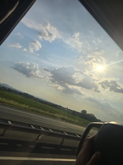 A photograph taken from within a moving vehicle captures a rural highway landscape under a partly cloudy sky, identified as being in the vicinity of Keşan, Turkey. The view is angled, with the dark frame of the vehicle's window visible on the left and top edges. The sun, emitting bright rays and lens flare, is prominently positioned in the upper right quadrant of the sky. Various cumulus and stratocumulus clouds are scattered across the blue-grey sky.

A multi-lane asphalt highway, marked with white lines, traverses the lower foreground diagonally from left to right, bordered by a metal guardrail. Lush green agricultural fields extend from the highway towards the mid-ground. Beyond the fields, a line of trees and scattered buildings, including a distant structure with a prominent spire or tower, lead to a hazy horizon featuring indistinct hills or mountains. A human hand, out of focus, is visible gripping a dark headrest or seat component in the lower right corner of the frame. The lighting conditions suggest either late afternoon or early morning.