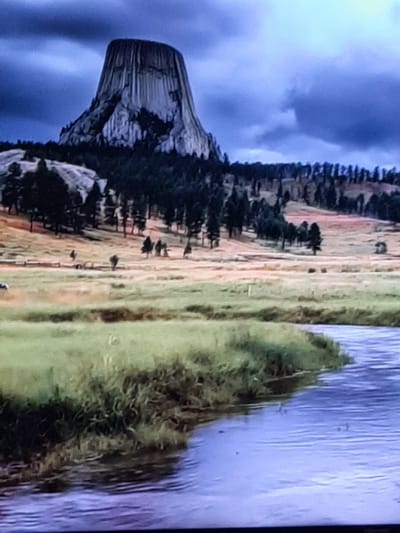 The image shows a landscape with a large, prominent rock formation on the left, resembling a tower or a natural monument. This rock is dark and appears to have a textured surface with vertical lines. It is surrounded by dark green trees, and in the background, there is a vast expanse of dry, brownish-yellow grass or fields, with some patches of green. The sky above is a dark, cloudy blue. To the right of the rock formation and trees, there is a body of water, likely a river or a lake, with a blue-purple hue and visible ripples on its surface. The lighting suggests it might be daytime, but the dark clouds create a somewhat dramatic or overcast atmosphere. The scene evokes a sense of natural grandeur and ruggedness. There is no text visible in the image.