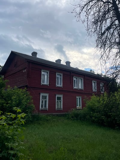The image shows a two-story, red wooden house with a dark roof and visible chimneys. The house has multiple windows with white frames. Green foliage, including trees and bushes, surrounds the house, and a grassy lawn is in the foreground. The sky is overcast with clouds. The house is located in the village of Bluzhski Selski Sovet, Belarus.