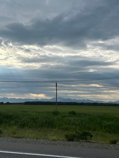 The image depicts a rural landscape under a cloudy sky with sunlight filtering through in some areas. In the foreground, a paved road with a white line is visible. Beyond the road is a green field with tall grass and some smaller plants. In the mid-ground, a dark line of trees stretches across the horizon, backed by a range of snow-capped mountains. A single wooden utility pole stands in the field, with wires stretching horizontally across the frame. The scene appears to be set in the city of Кеткино, Russia.