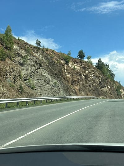 The image shows a roadside view of a steep, rocky hillside with scattered trees under a bright blue sky with scattered white clouds. A paved road with a white dividing line curves to the right in the foreground. A metal guardrail runs along the edge of the road, separating it from the grassy embankment leading up to the rocky slope. The hillside is layered with different rock strata and patches of dry soil and vegetation. Several coniferous and deciduous trees are growing on the hillside, some appearing young and small, others more mature. The lower part of the image shows the dashboard or hood of a vehicle. This scene is consistent with a natural landscape found along a road in a region like Усмангали, Russia, characterized by mountainous or hilly terrain.