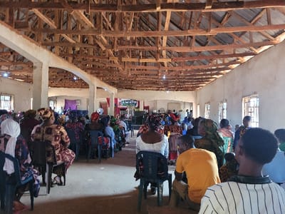 An indoor, wide-angle view depicts a large gathering within a functional, unadorned hall in Rinze, Nigeria. Approximately 50-70 individuals, primarily adults, are seated in rows on plastic chairs, facing a designated front area of the room. The majority are dressed in diverse, often brightly patterned traditional African attire, with many women wearing head coverings.
The environment features rough-textured, light-colored walls, likely plaster over concrete, exhibiting signs of wear and varying finishes. Multiple rectangular windows with metal security grilles are visible along the right wall and in the distant background, admitting natural light. The ceiling consists of exposed wooden roof trusses and purlins supporting a corrugated metal roof, with a few bare light bulbs suspended from the structure. Concrete pillars punctuate the space, supporting the roof. The floor is a light-colored, possibly concrete or dirt surface.
At the far end of the hall, a stage-like area is present. A red banner or screen with white text and graphics is visible on the right side of this area, along with what appears to be audio equipment, including speakers. A purple fabric or banner is draped on the wall behind the stage. One individual, a male in a white shirt, is visible standing near the stage. The attendees appear attentive, generally facing forward, indicating an ongoing event or service.
