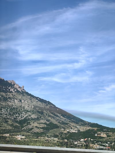 The image shows a mountainous landscape under a partly cloudy blue sky. The mountains are covered with sparse vegetation and rocky outcrops. In the mid-ground, there is a cluster of buildings, suggesting a settlement. A metal barrier is visible in the foreground, running horizontally across the bottom of the image. The scene appears to be a scenic view of the outskirts of Kiparisnoe, Ukraine, with the mountains forming the backdrop to the residential area.