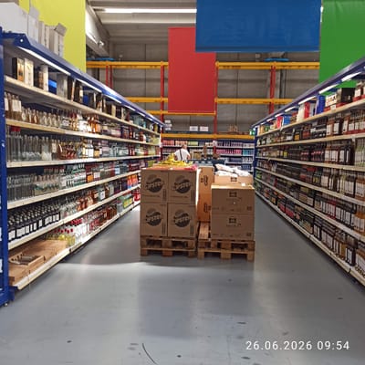 This media file captures an interior view of a retail store or warehouse, likely a supermarket, with rows of shelves stocked with beverages, predominantly bottles of alcohol and possibly other liquids. The perspective is from a high vantage point, looking down the aisles.
The scene is characterized by numerous shelves filled with a wide variety of products. The aisle on the right is particularly full, displaying many rows of bottles of different shapes, colors, and sizes, suggesting a diverse selection of alcoholic beverages. Some bottles are clear, others amber, and some contain darker liquids. The shelves themselves appear to be made of metal, with wooden accents on some.
In the foreground, several cardboard boxes are stacked on wooden pallets. Some of these boxes display text, including "CIPSTER," "SAN CARLO rustica," and "virtual." One box clearly states "12 multipack da 132g."
Towards the left side of the image, there are large, colored panels hanging from above – one green, one blue, and one red. These may be decorative elements or signage. In the background, industrial shelving and structures are visible, along with concrete walls and floors, indicative of a large commercial space. A person wearing a white shirt is partially visible on the left, leaning towards a shelf, suggesting they might be an employee or a shopper.
The timestamp "26.06.2026 09:54" is overlaid on the image, indicating the date and time of capture. The lighting suggests an indoor environment, possibly illuminated by overhead store lights, casting some shadows on the floor. The location context provided is Terlizzi, Italy.