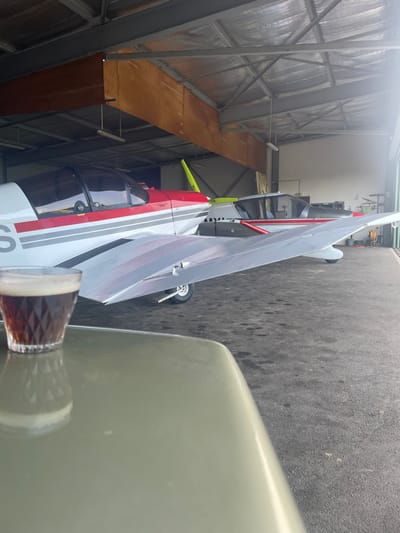The scene is set inside an aircraft hangar at Chonas-l'Amballan, France. In the immediate foreground, a faceted glass containing a dark brown liquid with a thin layer of off-white foam is positioned on a light olive-green, reflective surface, likely a table. Directly beyond this surface, the silver-grey wing of a small aircraft extends into the midground. The midground features two parked light aircraft. The primary aircraft, positioned to the left, is predominantly white with a prominent red stripe and a thinner grey stripe along its fuselage, a clear cockpit canopy, a visible yellow-tipped propeller, and a single nose landing gear wheel. Behind this, a second, darker grey aircraft with red accents is partially visible. The hangar exhibits a corrugated metal ceiling with silver insulation, exposed grey metal structural beams, and sections of light brown wooden paneling along the upper walls. Fluorescent light fixtures are suspended from the ceiling structure. The floor is a dark, textured asphalt or concrete surface. An open hangar door on the right side of the frame allows natural light to enter the space. No people are present.
