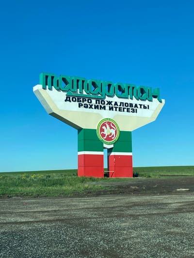 The image shows a sign for the city of Kamaevo, Russia, set against a clear blue sky. The sign is green on top, with the city's name in Russian and Tatar languages. Below the name, there is a section in beige, containing the phrase "ДОБРО ПОЖАЛОВАТЬ! РӘХИМ ИТЕГЕЗІ" (Welcome in Russian and Tatar). Below that, the sign is painted in the colors of the flag of Tatarstan: green, white, and red. On the white stripe, the coat of arms of Tatarstan is featured. The sign is situated next to a road, with gravel and grassy areas visible to the right.