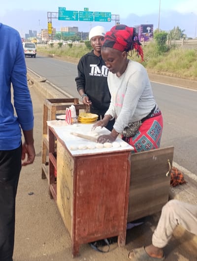 Two women stand next to a small wooden food cart on a roadside. One woman, wearing a red and black headscarf and a gray shirt, is rolling out dough on the cart. The second woman, wearing a white beanie and a black hoodie, looks on. A few other people are standing near the cart. A road and highway sign are in the background, with traffic and greenery visible beyond. The cart has writing on its side.