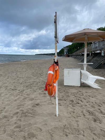 The image depicts a sandy beach in Pionersky, Russia, under a cloudy sky. In the foreground, a white pole stands with an orange life preserver hanging from it. Behind the pole, a white rectangular structure, likely a storage box, is visible, along with a stack of white plastic chairs and a large beige umbrella. A walkway with a metal railing runs along the edge of the beach, lined with trees and buildings in the background. The sea stretches out to the horizon. The scene suggests a beach that is either closed for the season or experiencing inclement weather, as indicated by the empty chairs and the overcast sky.