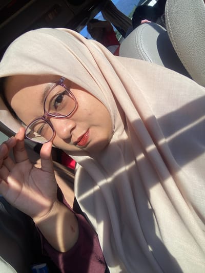 The image depicts an adult female seated inside a vehicle in Sumber Suko, Indonesia. She is positioned facing the camera, wearing a light beige fabric hijab that covers her head and drapes over her shoulder and chest. She wears purple-framed eyeglasses, which she holds with her left hand near the lower frame. Her lips are coated with a reddish-brown lip product. Her skin tone is light to medium, with a small, reddish mark visible on her left forearm/wrist. She is dressed in a reddish-brown garment. The car interior features light grey leather seats with visible stitching. Strong, direct sunlight enters the vehicle, casting defined linear shadows across her face, hijab, and arm, indicating a bright, outdoor daytime setting. In the background, another individual wearing a red top is partially visible and out of focus, seated behind a grey car headrest. Beyond the car's interior, an out-of-focus view of a bright sky and distant natural landscape can be seen through a window. A small, blue-wrapped item is visible near her left hand on a surface.