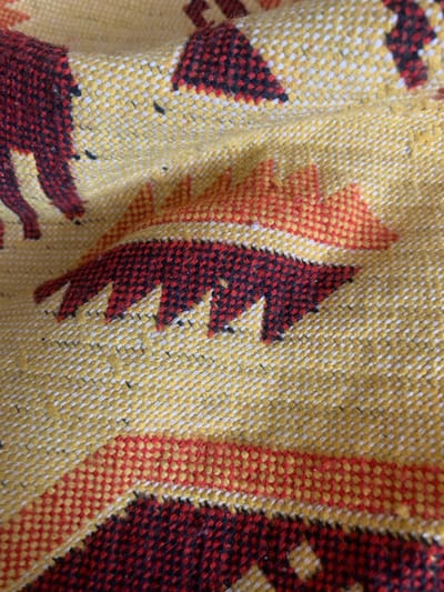 The image shows a close-up view of a colorful, woven textile, likely a blanket or rug, with a geometric pattern. The predominant colors are yellow, red, and black. The texture of the weave is clearly visible, with small, regular loops forming the pattern. There are abstract shapes and stylized animal figures in red and black against a yellow background, bordered by red and white stripes. The textile appears to be folded or creased in places, suggesting it is a soft material. The lighting is even, and the focus is sharp, highlighting the details of the fabric's construction and design. The image does not contain any elements that would allow for its location to be identified as being in Kovrov, Russia, or any other specific geographic location.