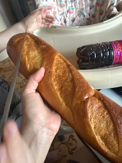 The image shows a close-up of a person's hands holding a baguette. The baguette is golden brown and appears to be fresh. A bottle of dark liquid, possibly a beverage, is visible in the background, with a label that says "ENDI". The setting seems to be indoors, with a patterned blanket or fabric visible on the left side of the frame. The lighting suggests it could be daytime. The focus is on the baguette and the hands, implying an activity related to preparing or eating it.
