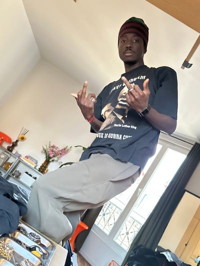 A person stands inside a building. The person is wearing a black t-shirt with an image of Martin Luther King Jr. and khaki pants, and a hat. The person is making hand gestures. In the background, there is a window and a shelf with flowers and other objects. The image was taken in Saint-Ouen-sur-Seine, France.