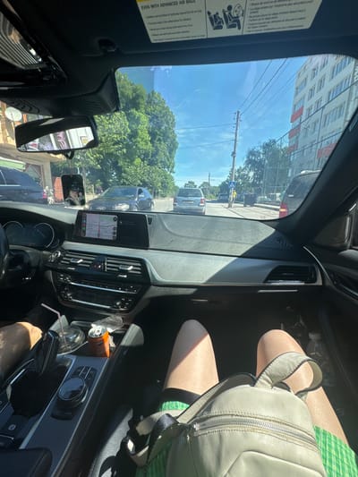 The image, taken from inside a car, shows an urban street scene in Byala Slatina, Bulgaria. The street includes buildings, trees, telephone poles, and parked cars. Inside the car, a backpack rests on the passenger's lap. On the car's dashboard, a can, a cup with a straw, and a phone displaying a map are visible. The car appears to be stopped in traffic.