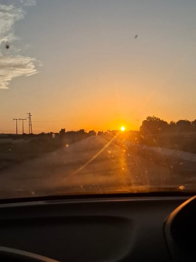 The image depicts a sunset scene viewed from inside a vehicle, likely on a road leading towards or away from the city of Gorna Malina, Bulgaria. The sun is a bright, low orb on the horizon, casting a warm, orange glow across the sky and reflecting off the road ahead. The sky transitions from a pale blue at the top to a vibrant orange closer to the horizon. Wispy clouds are visible on the left side of the frame. In the middle ground, power lines and utility poles stand silhouetted against the sky. Trees and vegetation line the sides of the road, also appearing as dark silhouettes. The foreground shows the interior of a car, with the dashboard and windshield visible. The windshield has some specks or marks on it, which create bokeh effects in the light. The overall impression is that of a peaceful, scenic drive during golden hour.