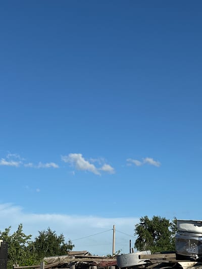 The media file shows a bright, clear blue sky with a few wispy white clouds on the right side. The image is oriented vertically, with the sky occupying the majority of the frame. On the far right, there is a section of what appears to be an outdoor junkyard or storage area. This area includes wooden structures, metal objects that resemble parts of appliances, and lush green trees. A wooden utility pole with wires is also visible, suggesting an inhabited area. The lighting indicates it is daytime, likely mid-morning to afternoon, given the brightness and the clear sky. There are no people or animals visible. The overall impression is one of an outdoor, possibly rural or semi-industrial setting on a sunny day. There is no visible text in the image. The location context provided is Tobyl, Kazakhstan.