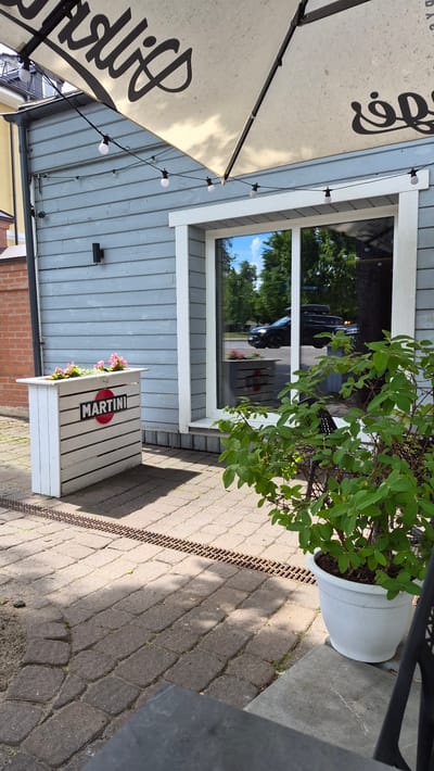 The image depicts the exterior of a building, likely a cafe or restaurant, with a light blue wooden facade. A large white umbrella with black lettering is partially visible at the top, along with a string of white lights. In front of the building, to the left, is a white wooden planter box adorned with a circular red and black "MARTINI" logo and filled with pink flowers. The ground is paved with grey rectangular stones. A metal grate runs across the paving stones. To the right, a large window reflects the environment, showing trees and a dark-colored car with a roof rack. A potted green plant in a white pot is situated to the right of the window. The overall scene is set outdoors, suggesting a sunny day. The architectural style and the presence of the Martini logo hint at a European setting. Although the precise location in Zarasai, Lithuania is not explicitly stated, the image captures a common urban outdoor dining or seating area.