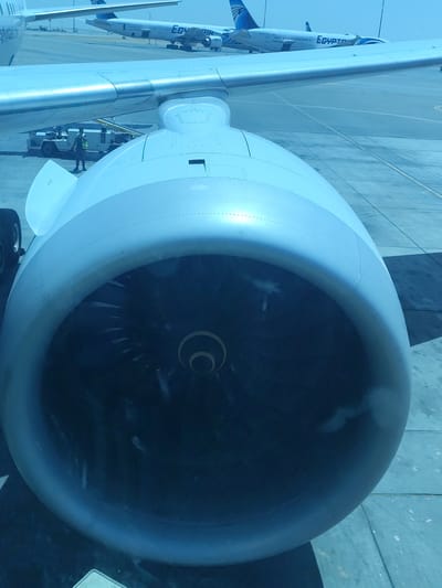The image provides a bright, somewhat cool-toned view from an airplane window, primarily focusing on the large turbofan engine of the aircraft the photo was taken from. The engine, painted in shades of light blue and grey, dominates the foreground, with its intricate fan blades visible inside the intake. Above and to the left of the engine, a portion of the aircraft's wing extends, showing rivets and faint "NO STEP" text on its surface.
In the background, under a clear, sunny sky, an airport tarmac is visible. Two other airplanes, both identified as "EgyptAir" aircraft by the prominent branding on their tails and fuselages (in English and Arabic script), are parked on the concrete surface. One of these background planes appears to be a narrow-body jet.
Under the wing of the foreground aircraft, a ground crew member, wearing a high-visibility vest, stands near a white ground service vehicle or equipment. The strong daylight suggests it is daytime, with clear weather conditions. The visible "EgyptAir" branding on multiple aircraft, combined with the location context, places the scene at an airport in Cairo, Egypt. The image captures a moment of ground operations or waiting at a gate.