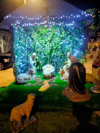 A nativity scene is depicted at night, illuminated by string lights. The scene features figurines of Mary, Joseph, baby Jesus in a manger, and various animals including a donkey, cows, and sheep. The figurines are arranged on a green surface mimicking grass, and the backdrop is adorned with green foliage. The structure overhead is simple, resembling a makeshift shelter. Additional figures stand to the side. A car is partially visible in the background.