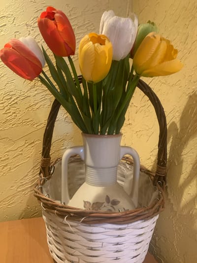 A still life image depicts a decorative arrangement of artificial tulips in a white woven basket. The basket contains a white ceramic vase with handles, adorned with a floral transfer print. The tulips are in various colors, including red, orange, yellow, white, and green. The basket and vase are placed on a wooden surface against a textured yellow wall. The scene is illuminated by overhead lighting, casting soft shadows. The image does not depict any people or specific actions. The location is not ascertainable from the visual information provided.