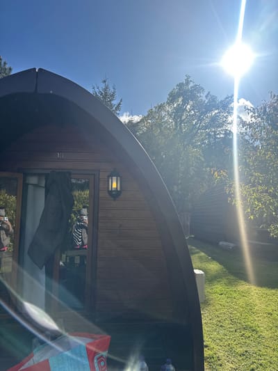 The image shows a wooden cabin with a number '10' on it. The cabin is located in a wooded area with a grassy lawn in front of it. The sun is shining brightly in the sky, creating lens flares. The cabin has a window with a jacket hanging on a hook and two people can be seen reflected in the glass. The cabin is situated in Cloughton, United Kingdom, at the coordinates 54.369 latitude and -0.506 longitude. There are two water bottles and a red and white bag on the ground in front of the cabin. There are trees in the background, a second, identical cabin, and a small, white structure. The sky is blue with white clouds.