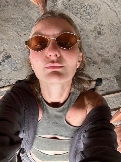 A light-skinned person with sunglasses is photographed from a low angle in what appears to be a rustic setting. The person is wearing a gray sports bra, partially covered by a dark gray hoodie or jacket. A tattoo is visible on their right shoulder. The person is sitting on a chair with wooden and metal components. The background consists of a rough, concrete floor with visible cracks and textures. The city of Yên Minh Commune, Vietnam, is not discernable in this scene.